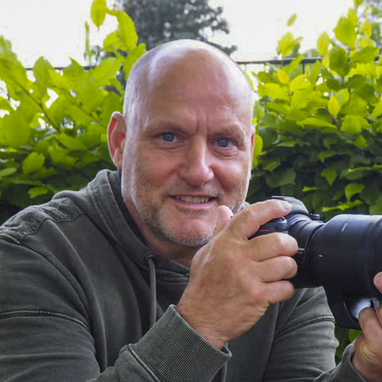 Der Fotograf Stefan Meier mit Kamera lächelt vor einer grünen Pflanzenkulisse.