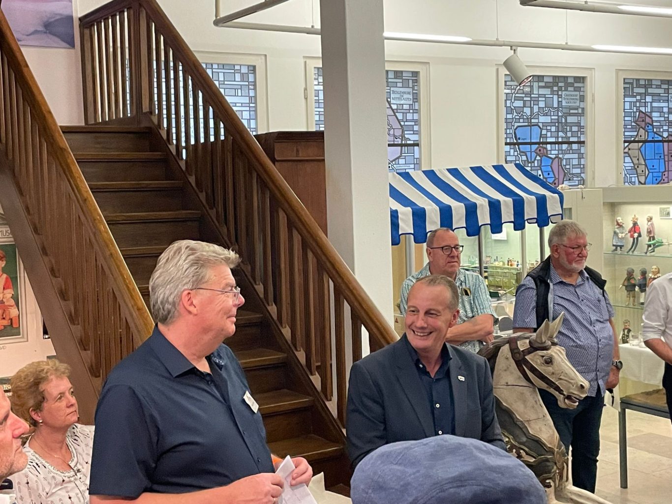 Gruppengespräch in einem Innenraum, ein Mann spricht, eine Holzpferd-Ruch daneben.